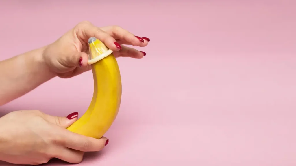 A imagem mostra apenas as mãos de uma mulher vestindo uma camisinha em uma banana.
