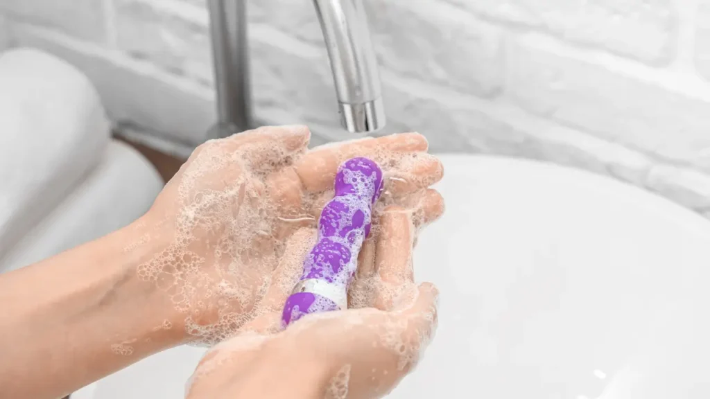 A imagem mostra uma pessoa lavando um vibrador na pia, com água e sabão.