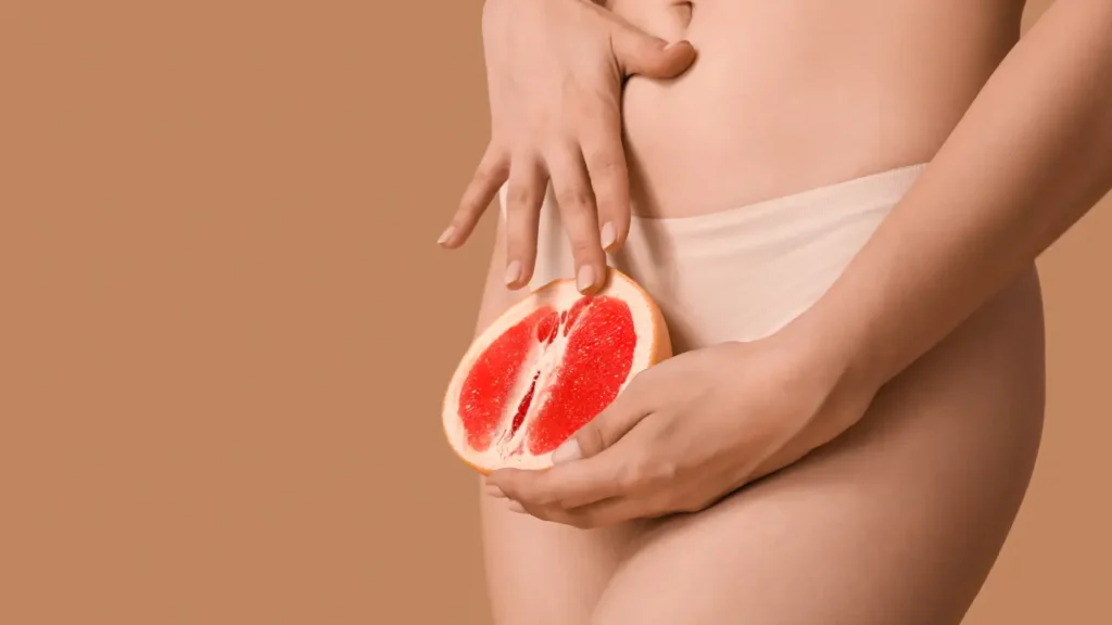 Mulher segurando uma tangerina partida ao meio na altura da pelvis, remetendo a como é o orgasmo feminino.